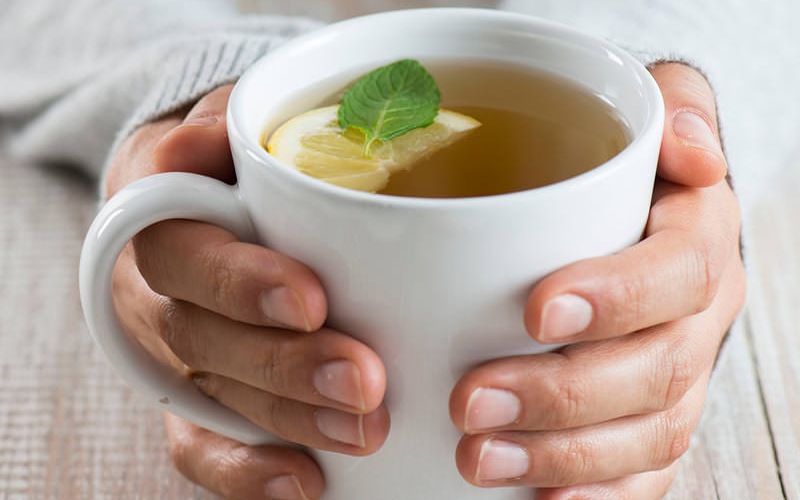 woman's hands hold a cup of tea with a slice of lemon and mint leaf floating in it