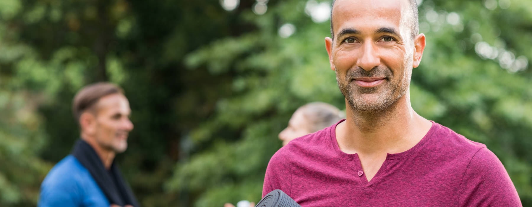 smiling man holds yoga mat in park with friends talking behind him